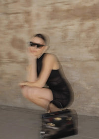 Person in a black dress sitting on a suitcase against a beige wall. #color_black